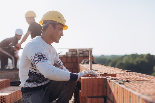 Construction worker standing on scaffolding in July sunlight, wearing safety gear, with a hazy blue sky and industrial background.